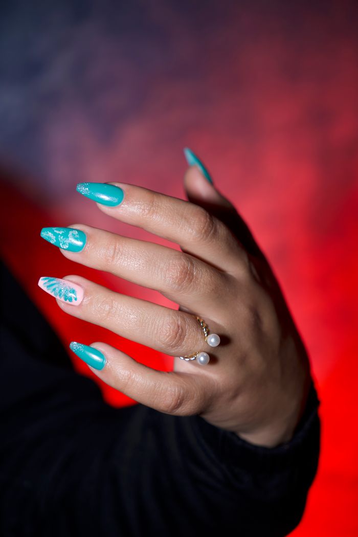 Close-up of a hand with turquoise nail art, adorned with rings and pearls, on a vibrant background.